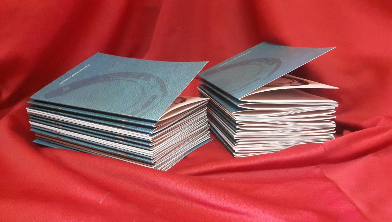 Dwa stosy broszur z niebiesko-zielonymi okładkami i wzorem okręgu na czerwonym, drapowanym tle.