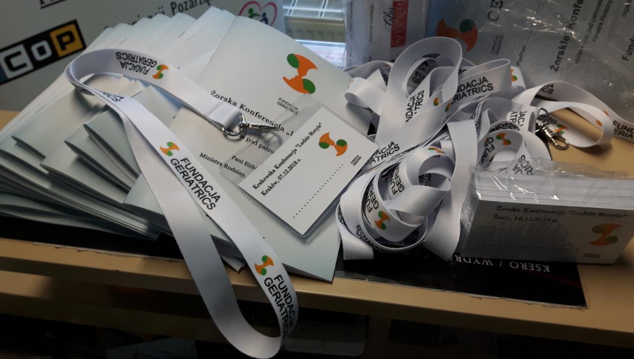 Stos ulotek i identyfikatorów konferencyjnych z logo Fundacji Geriatrics, widoczne smycze z nadrukiem nazwy fundacji i logo.