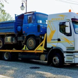 Niebieska ciężarówka Tatra przewożona na żółtej lawecie Renault TT Trans z numerem telefonu 518 636 015, w tle budynek z reklamą restauracji Bohema i baner wyborczy.