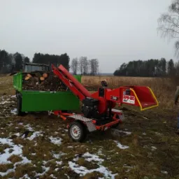Czerwona rębarka do gałęzi marki Remet RPS-120 podłączona do zielonego traktora z przyczepą pełną pociętych gałęzi na zaśnieżonym polu.