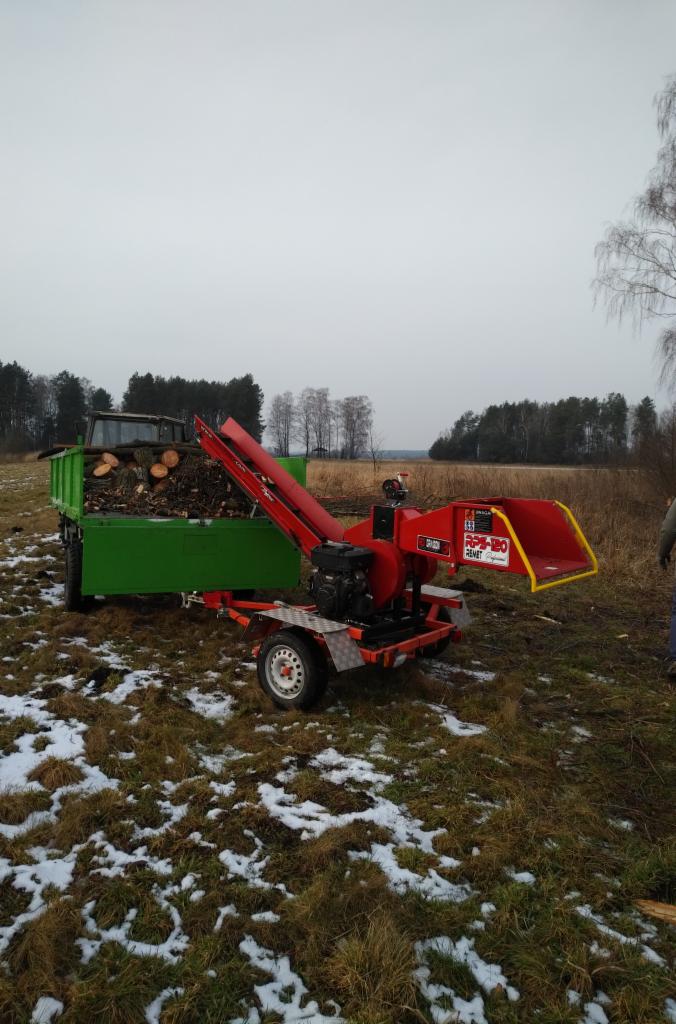 Czerwona rębarka do gałęzi marki Remet RPS-120 podłączona do zielonego traktora z przyczepą pełną pociętych gałęzi na zaśnieżonym polu.