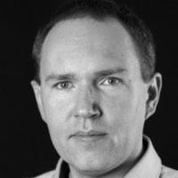 Portret biznesowy mężczyzny w czerni i bieli, z wyraźnymi rysami twarzy i krótkimi włosami, na ciemnym tle. Ujęcie od przodu, skupione na twarzy.