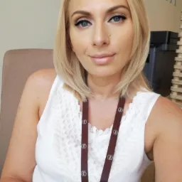 Portret kobiety z blond włosami i identyfikatorem na szyi, w tle biuro z drukarką i ścianą z napisem.