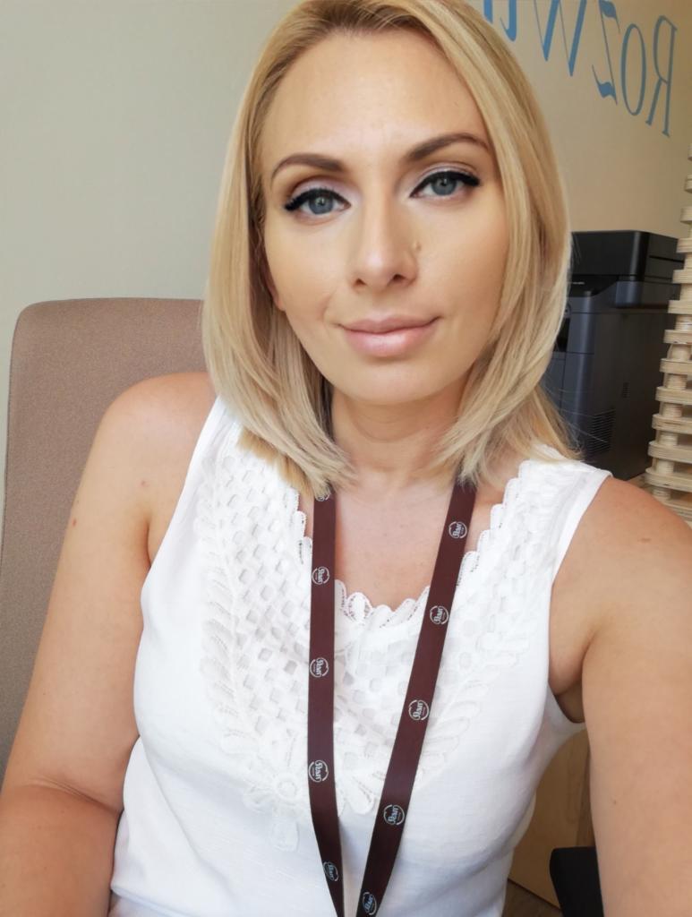 Portret kobiety z blond włosami i identyfikatorem na szyi, w tle biuro z drukarką i ścianą z napisem.