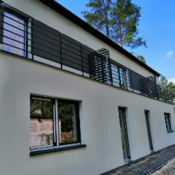 Nowoczesny dom z balkonem zabezpieczonym metalową, poziomą balustradą w kolorze grafitowym, widok z zewnątrz, kostka brukowa ułożona wzdłuż ściany budynku.