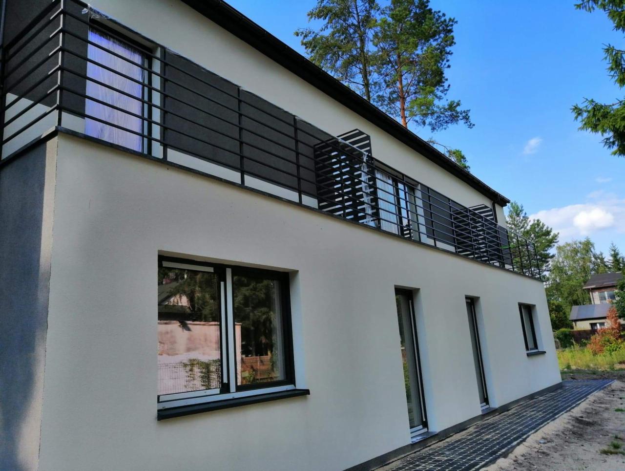 Nowoczesny dom z balkonem zabezpieczonym metalową, poziomą balustradą w kolorze grafitowym, widok z zewnątrz, kostka brukowa ułożona wzdłuż ściany budynku.