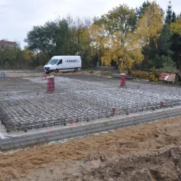 Zbrojenie fundamentów pod budynek, widoczne stalowe pręty tworzące siatkę, szalunek z betonu, na tle krajobraz z drzewami i budynkiem oraz biały samochód dostawczy firmy.