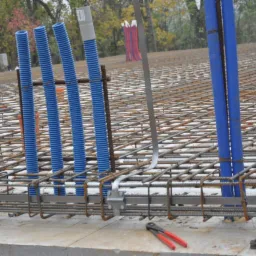 Siatka zbrojeniowa z pionowymi niebieskimi rurami karbowanymi i czerwonymi rurami w tle, widoczne narzędzia do cięcia drutu.