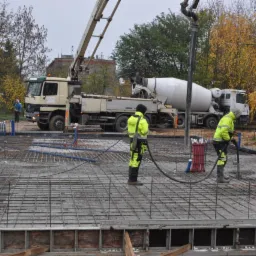 Wylewanie betonu na zbrojenie fundamentów z użyciem pompy do betonu, pracownicy w odblaskowych ubraniach, widoczne przewody instalacyjne w kolorach niebieskim i czerwonym.