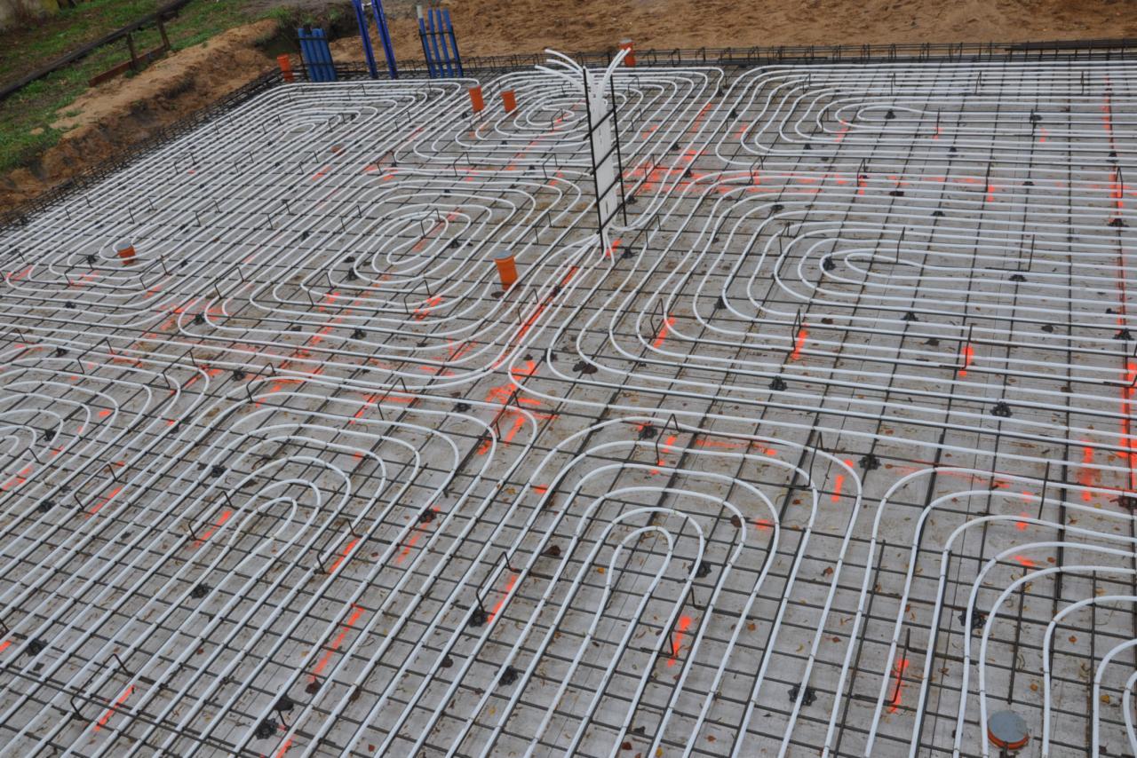 Siatka zbrojeniowa z rurami ogrzewania podłogowego ułożona na betonie, pomarańczowe znaczniki, widok z góry.