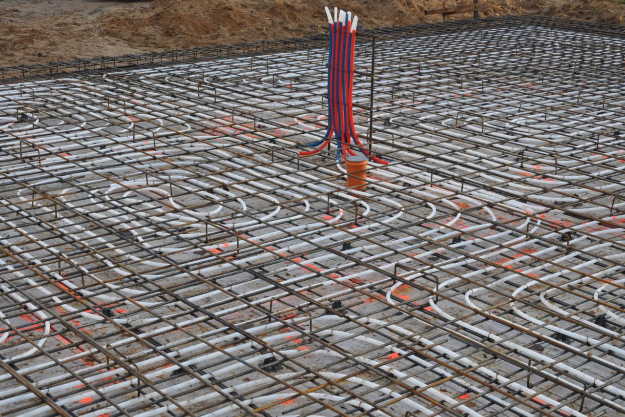 Siatka zbrojeniowa z rurami ogrzewania podłogowego ułożona na przygotowanym podłożu, widoczne pionowe rury instalacyjne w kolorze czerwonym i niebieskim.