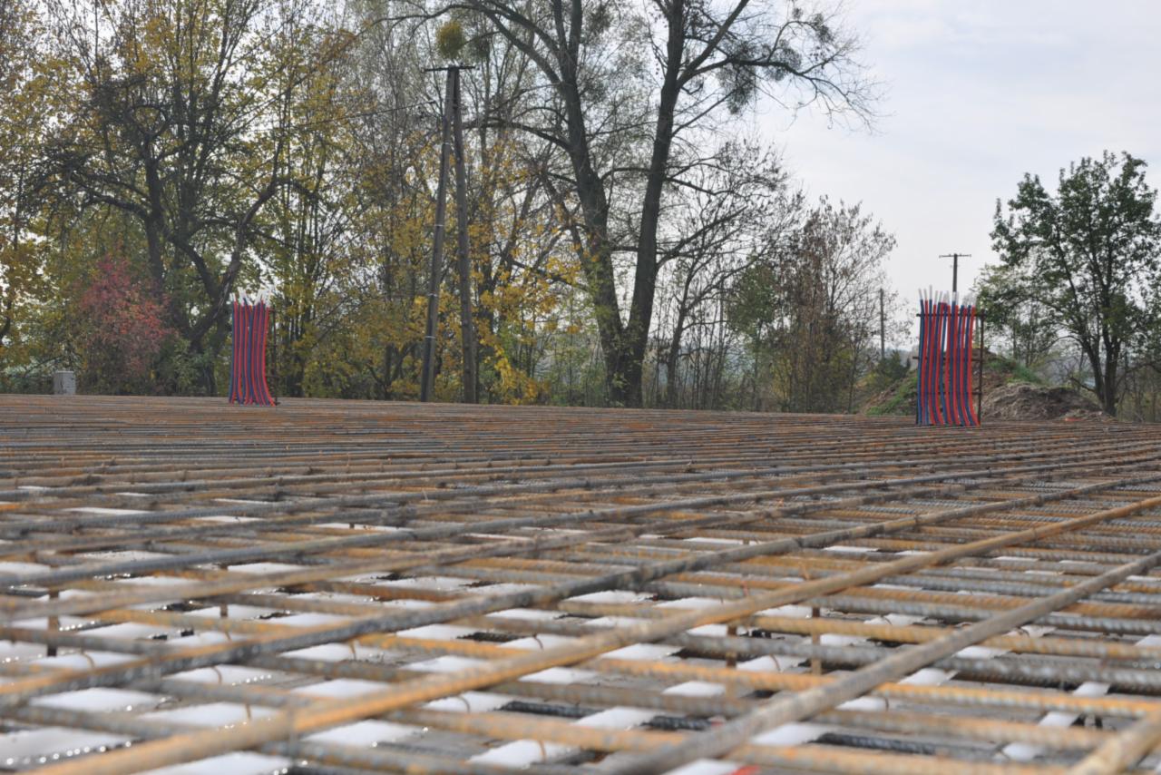 Siatka zbrojeniowa na przygotowanym podłożu, widoczne pionowe rury w czerwono-niebieskiej izolacji, drzewa w tle.