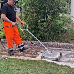 Mycie u Siwego - Pracownik w pomarańczowych spodniach czyści kostkę brukową myjką ciśnieniową. Widoczne piana i czyszczona powierzchnia. Tło: zieleń, krzewy, fragment budynku.