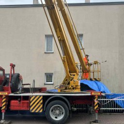 Mycie u Siwego - Pracownik w pomarańczowym kombinezonie na podnośniku koszowym czyści elewację budynku. Widoczny fragment elewacji, podnośnik z platformą i czerwona ciężarówka.