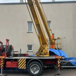 Pracownik w pomarańczowym kombinezonie na podnośniku koszowym czyści elewację budynku. Widoczny fragment elewacji, podnośnik z platformą i czerwona ciężarówka.