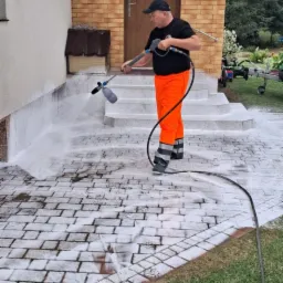 Pracownik w pomarańczowych spodniach czyści kostkę brukową myjką ciśnieniową, piana pokrywa powierzchnię. Budynek i trawnik w tle.