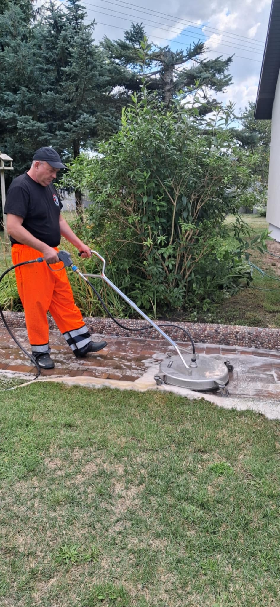 Pracownik w pomarańczowych spodniach czyści kostkę brukową myjką ciśnieniową. Widoczne piana i czyszczona powierzchnia. Tło: zieleń, krzewy, fragment budynku.