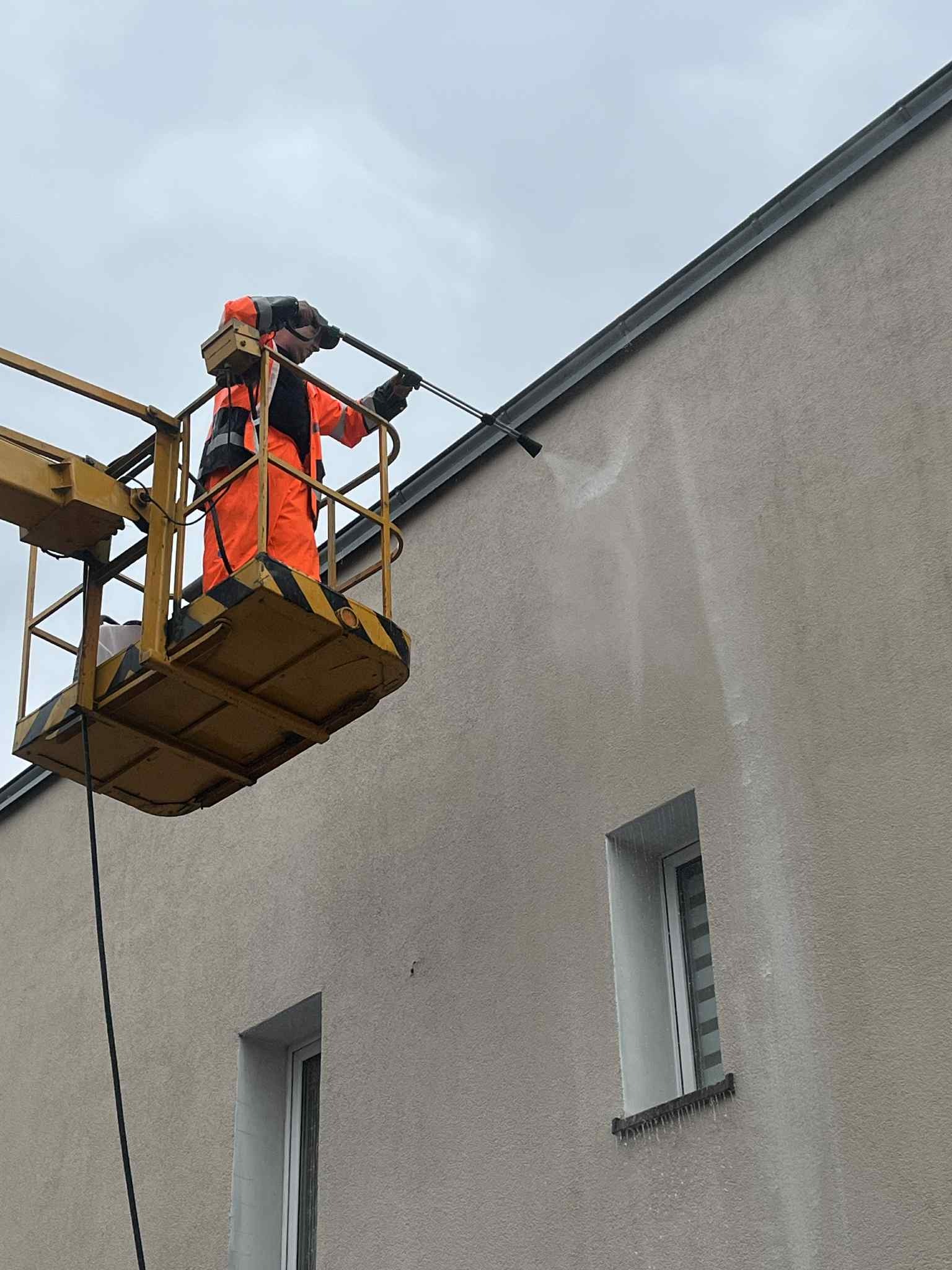 Pracownik w pomarańczowym kombinezonie na podnośniku czyści elewację budynku za pomocą myjki ciśnieniowej. Woda spływa po elewacji i szybach okiennych.