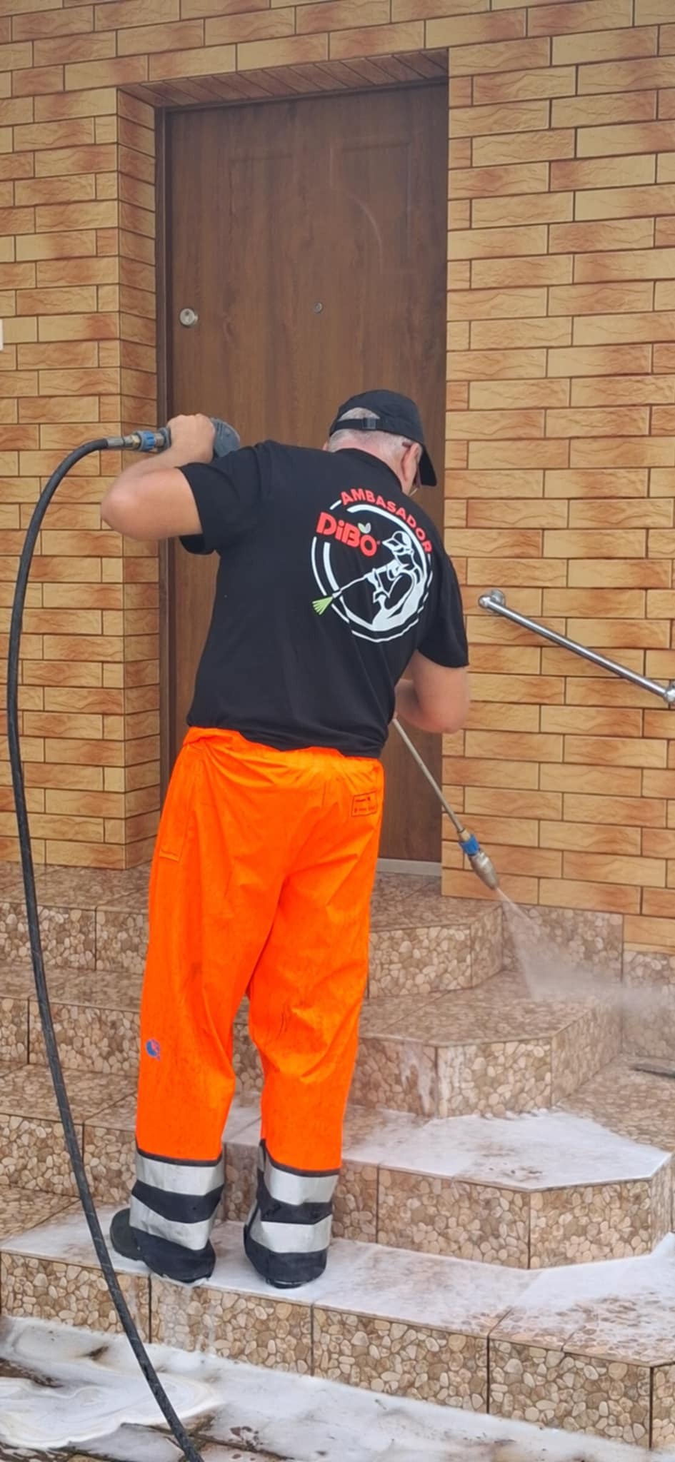 Pracownik w stroju roboczym czyści schody wejściowe do domu za pomocą myjki ciśnieniowej. Widoczna elewacja budynku z cegły.