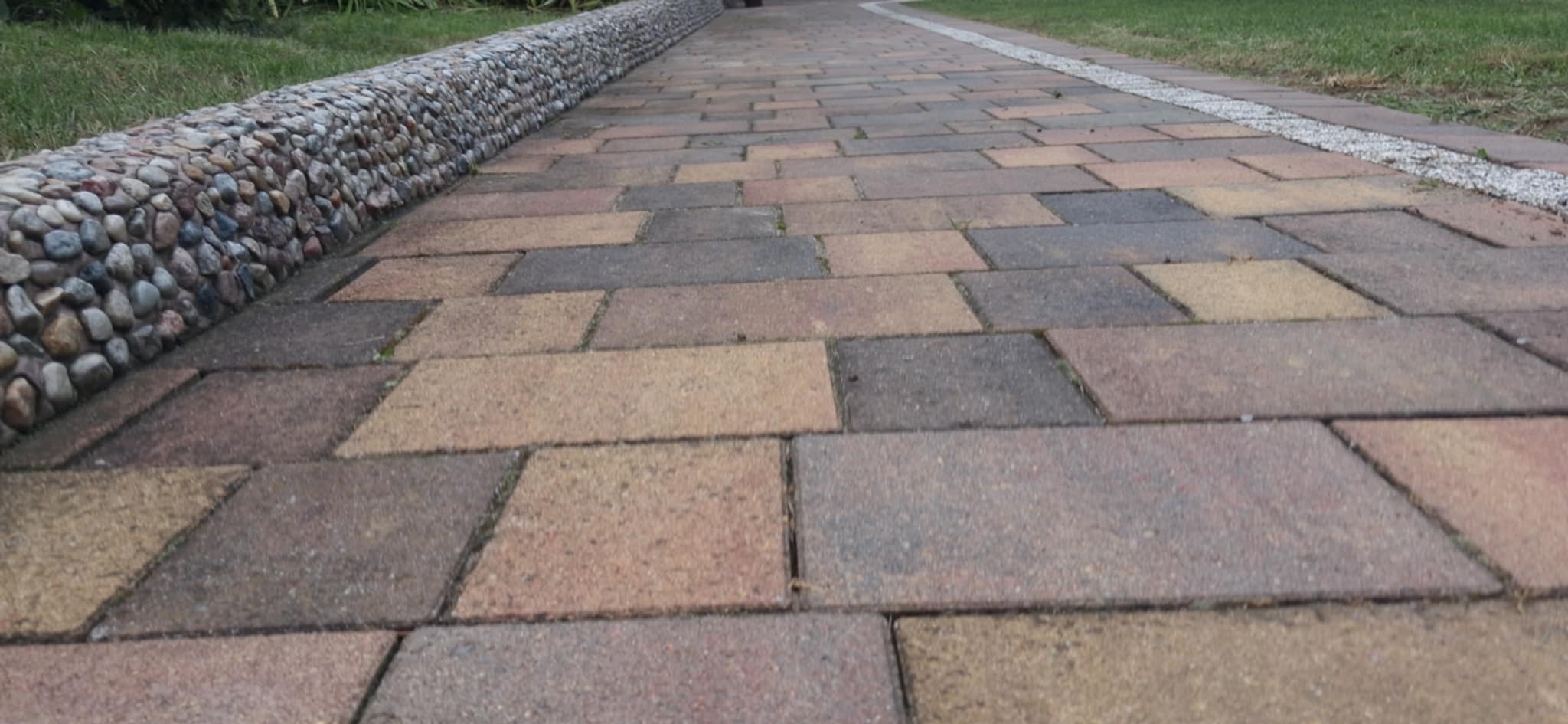 Ułożona kostka brukowa w odcieniach brązu i szarości, z ozdobnym obrzeżem z kamieni i żwiru, otoczona zielenią trawnika. Widok z perspektywy chodnika.