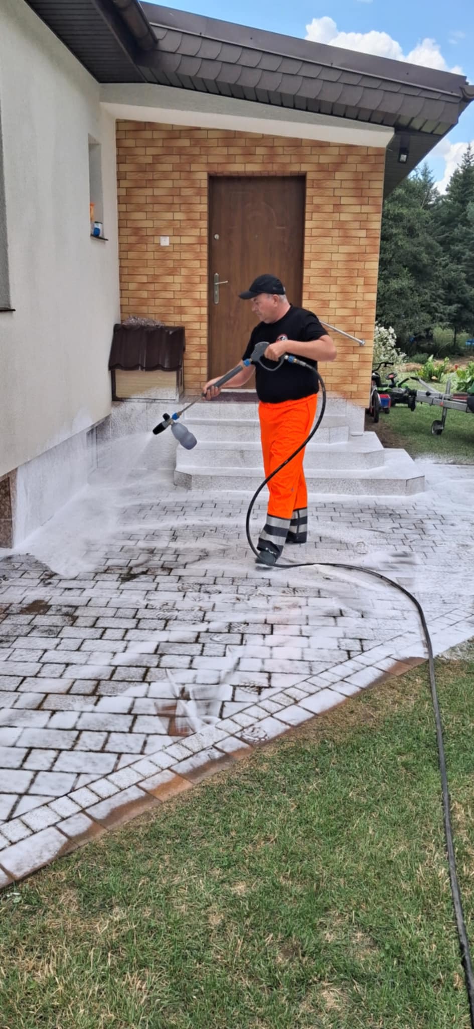 Pracownik w pomarańczowych spodniach czyści kostkę brukową myjką ciśnieniową, piana pokrywa powierzchnię. Budynek i trawnik w tle.
