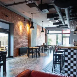 Wnętrze lokalu usługowego w Warszawie z ceglaną ścianą, oświetleniem przemysłowym i barem z czarno-białą szachownicą, widoczne stoliki i krzesła.