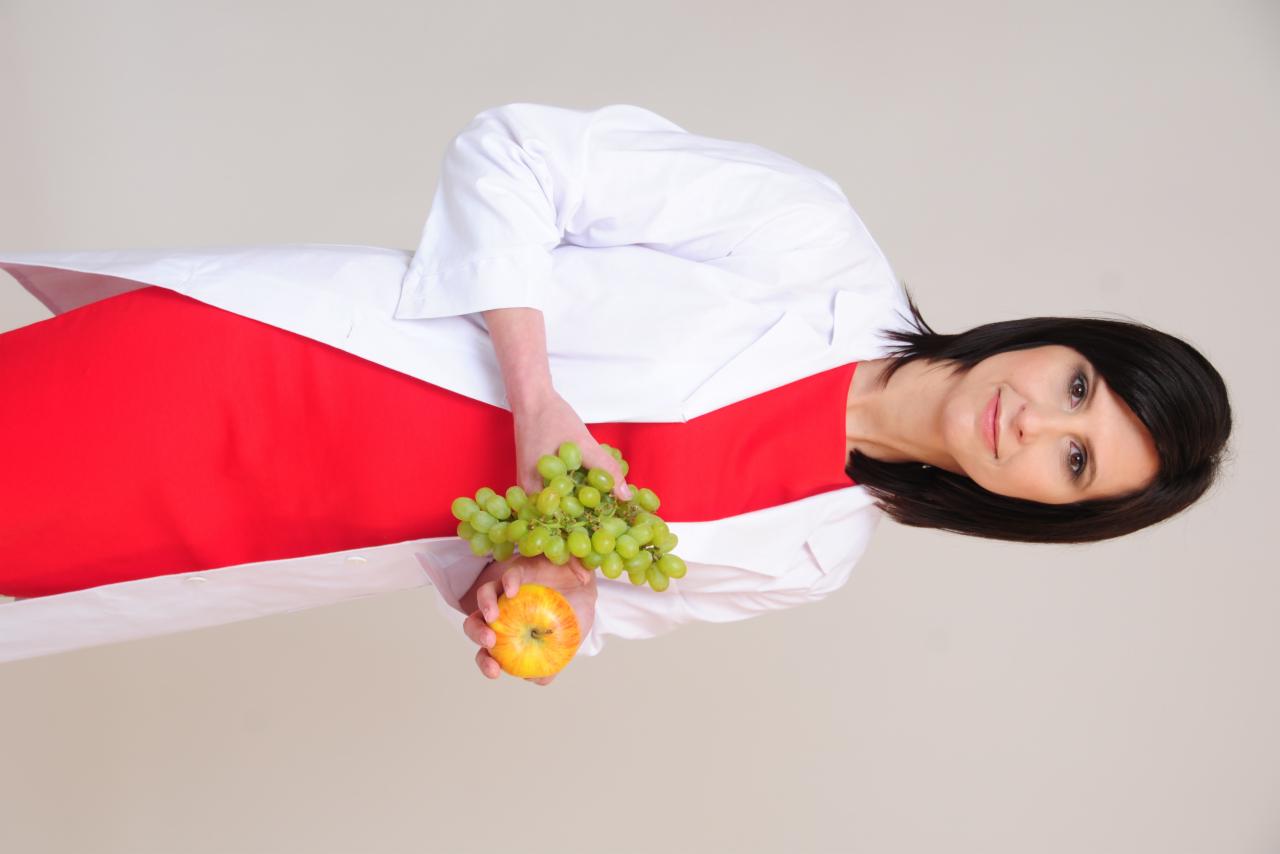 Uśmiechnięta kobieta w białym fartuchu trzyma jabłko i winogrona, prezentując zdrowe odżywianie. Czerwona sukienka dodaje kontrastu.