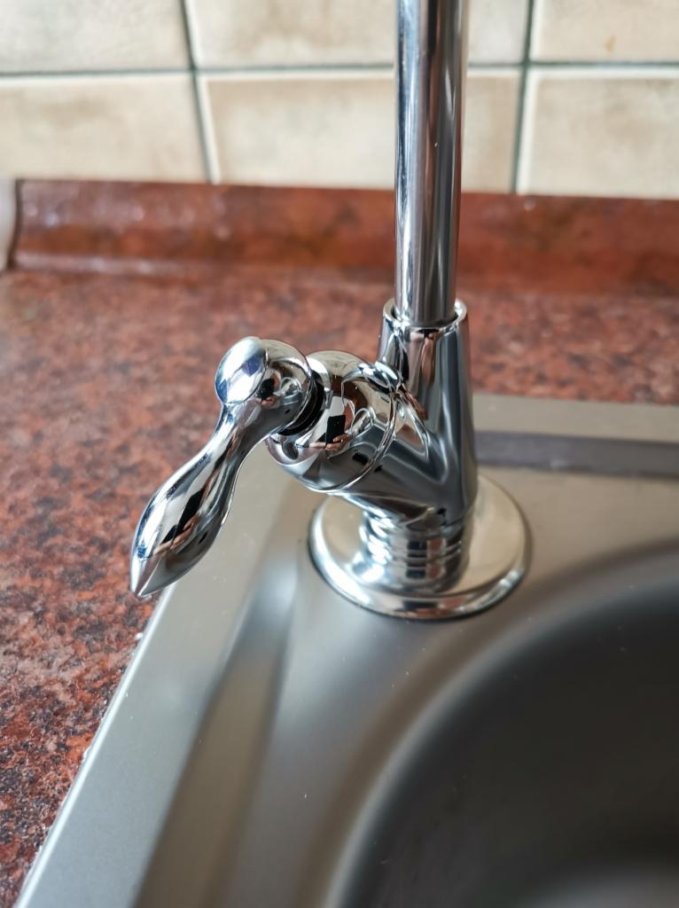 Błyszcząca, chromowana bateria kuchenna z pojedynczą dźwignią, zamontowana w metalowym zlewie, na tle płytek ściennych w odcieniach beżu i brązu.