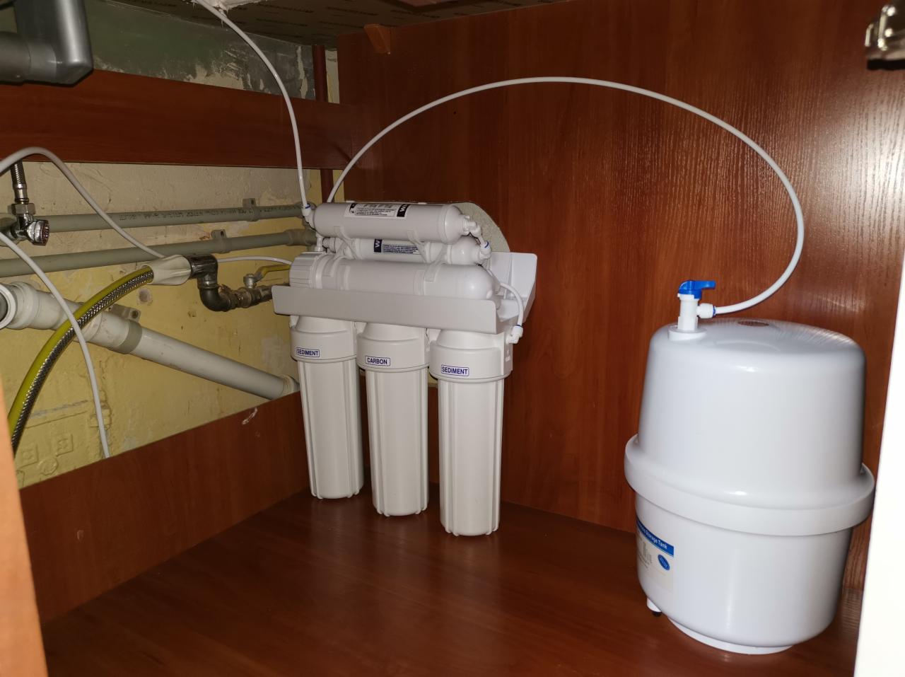 Zamontowany system filtracji wody pod zlewem z trzema filtrami wstępnymi oznaczonymi jako 'Sediment' i 'Carbon' oraz zbiornikiem na wodę z niebieskim zaworem.