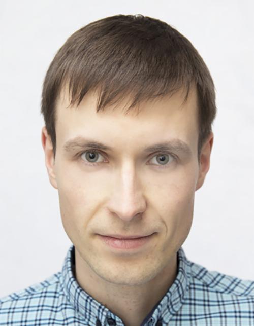 Portret młodego mężczyzny z krótkimi, brązowymi włosami, ubranego w niebieską koszulę w kratę, na jasnym tle. Uśmiech, naturalny wyraz twarzy.