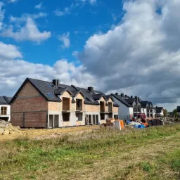 Szeregówka domów w trakcie budowy, widoczne ściany z pustaków, czarne dachy, drabina oparta o ścianę, niebo z chmurami w tle.