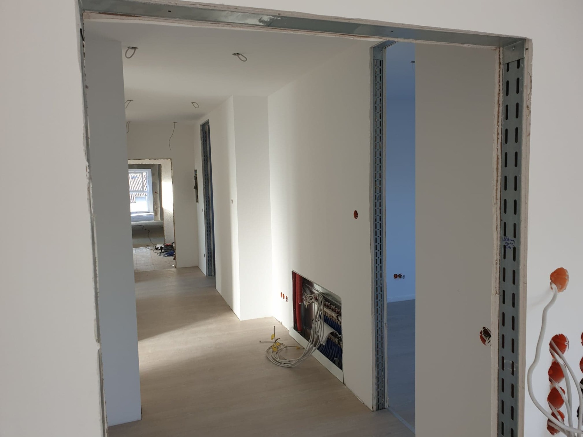 Wnętrze w trakcie wykańczania: widok na korytarz z metalowymi profilami przy otworach drzwiowych, instalacja elektryczna w ścianie i przewody na podłodze.
