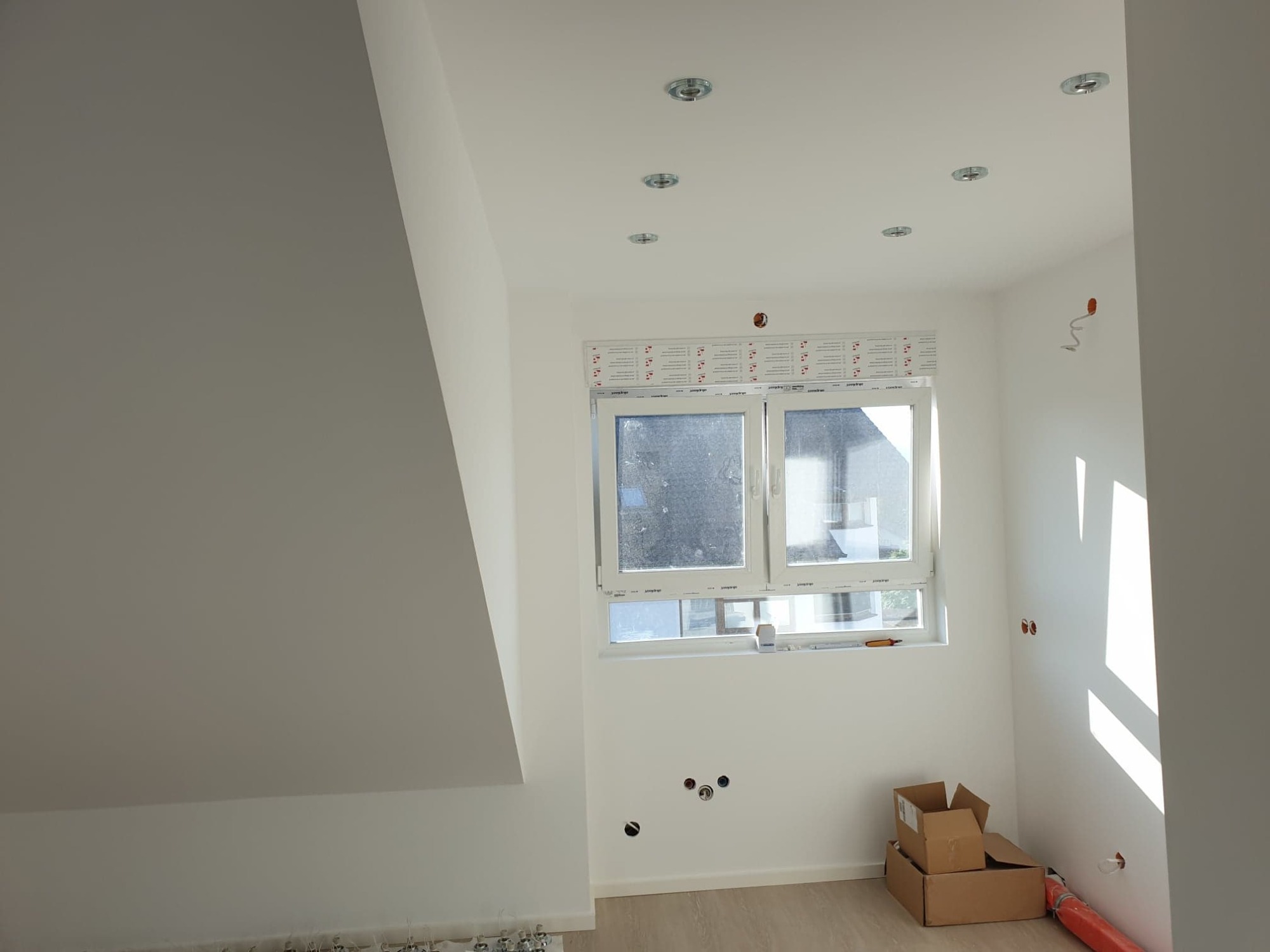 Wnętrze poddasza w trakcie wykańczania: okno z naklejoną folią ochronną, przygotowane punkty oświetleniowe w suficie, instalacja elektryczna w ścianie, kartony na podłodze i elementy instalacji...
