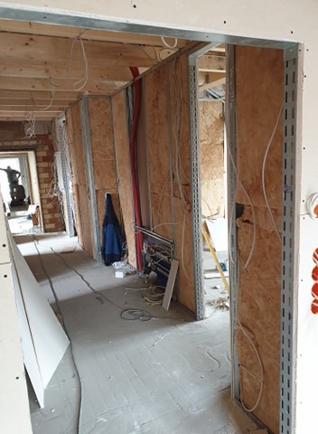 Wnętrze domu w trakcie wykańczania: widoczne ściany z płyt OSB, metalowe profile, instalacja elektryczna w peszlach, surowa podłoga betonowa i fragmenty płyt kartonowo-gipsowych.