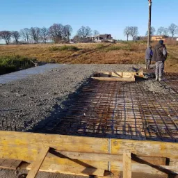 Wylewanie betonu na zbrojenie w drewnianym szalunku z widocznym betonowozem i pompą do betonu w tle.