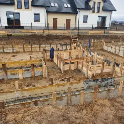 Wykop pod fundamenty z drewnianym szalunkiem i zbrojeniem stalowym, widok z g&oacute;ry na tle budowanego domu jednorodzinnego z białymi ścianami i ciemną dach&oacute;wką.