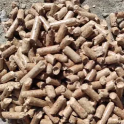 Stos brązowych, cylindrycznych peletek z widocznymi śladami trocin na szarym, betonowym podłożu.