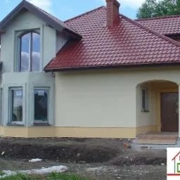 Nowo wybudowany dom jednorodzinny z czerwoną dachówką, kremową elewacją i widocznymi śladami prac ziemnych wokół budynku.