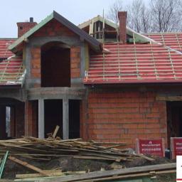 Daneko - Dom w trakcie budowy z czerwonej cegły Porotherm, widoczna konstrukcja dachu z czerwonymi dachówkami i drewnianymi łatami, niezamontowane okna i drzwi, palety z cegłami Porotherm na pierwszym...