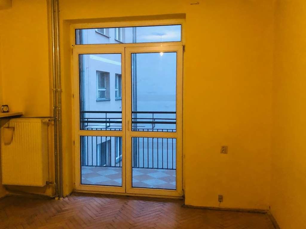 Żółte wnętrze pokoju z grzejnikiem i balkonem z widokiem na budynek naprzeciwko.