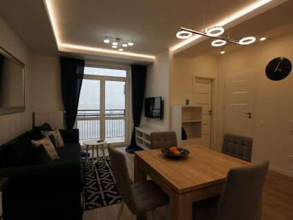 Jasne, nowocześnie urządzone wnętrze salonu z balkonem, drewnianym stołem i designerskim oświetleniem sufitowym.