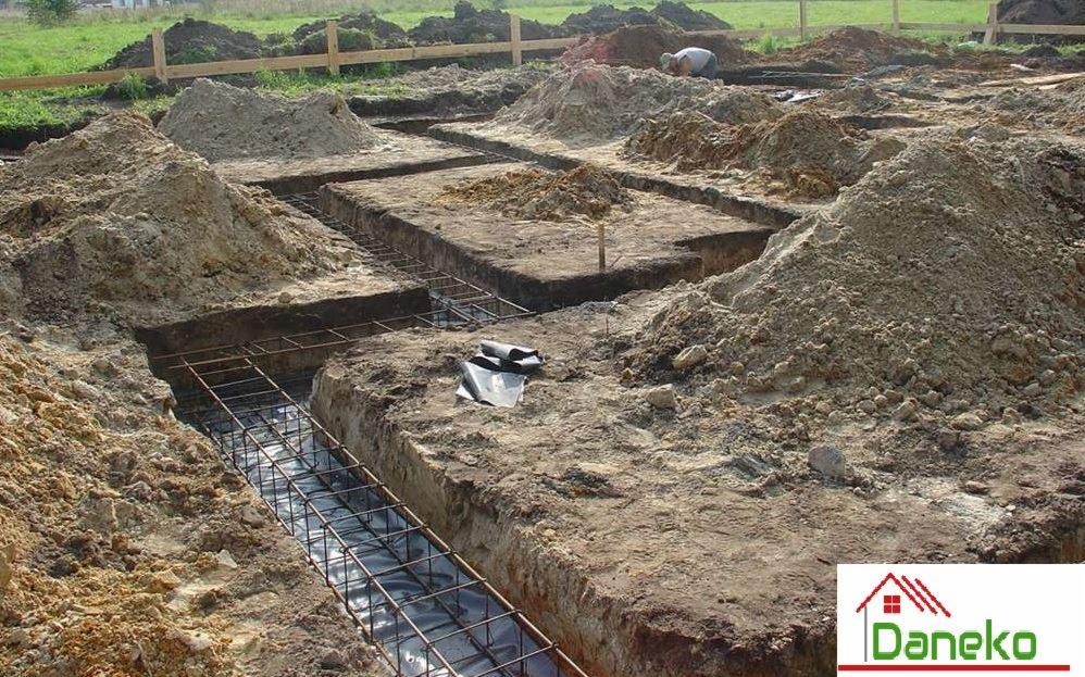 Wykonane wykopy pod fundamenty z widocznym zbrojeniem w rowach, folią izolacyjną i pryzmami ziemi, w tle częściowo widoczna osoba pracująca na budowie.