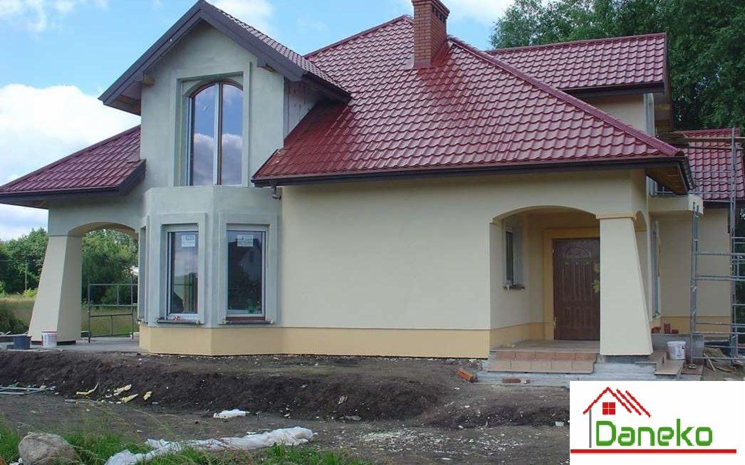 Nowo wybudowany dom jednorodzinny z czerwoną dachówką, kremową elewacją i widocznymi śladami prac ziemnych wokół budynku.