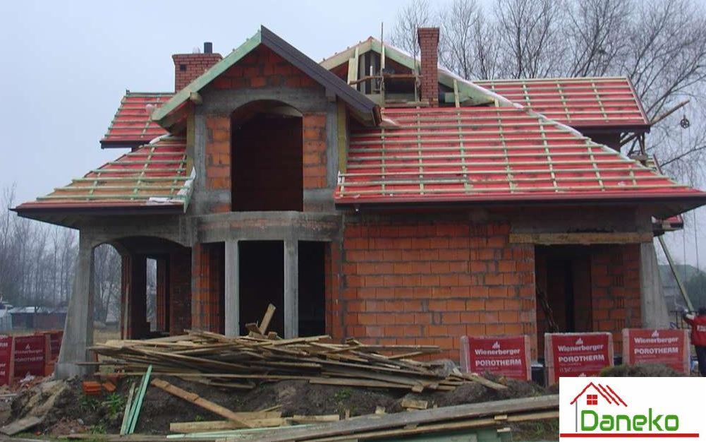 Dom w trakcie budowy z czerwonej cegły Porotherm, widoczna konstrukcja dachu z czerwonymi dachówkami i drewnianymi łatami, niezamontowane okna i drzwi, palety z cegłami Porotherm na pierwszym...