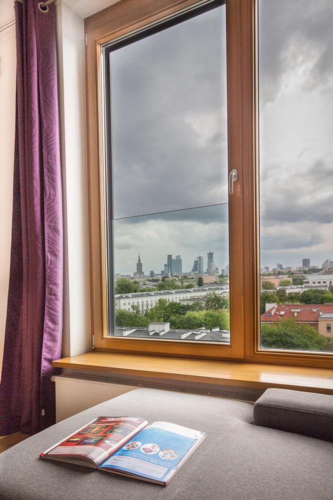 Nowoczesne okno z widokiem na panoramę miasta w pochmurny dzień, fioletowa zasłona po lewej stronie, otwarta broszura na szarym siedzisku.