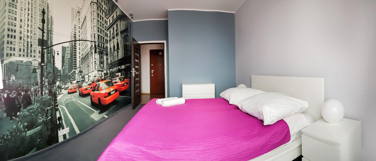 Pokój z fototapetą przedstawiającą ulicę Nowego Jorku z żółtymi taksówkami, łóżko z różową narzutą i białą pościelą, białe szafki nocne i komoda.