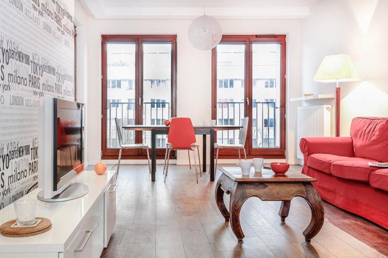 Jasne wnętrze mieszkania z drewnianą podłogą, czerwonymi akcentami w postaci sofy i krzesła, stół z owocami, widok na balkon przez okna z drewnianą ramą, oraz ściana z motywem napisu miast.