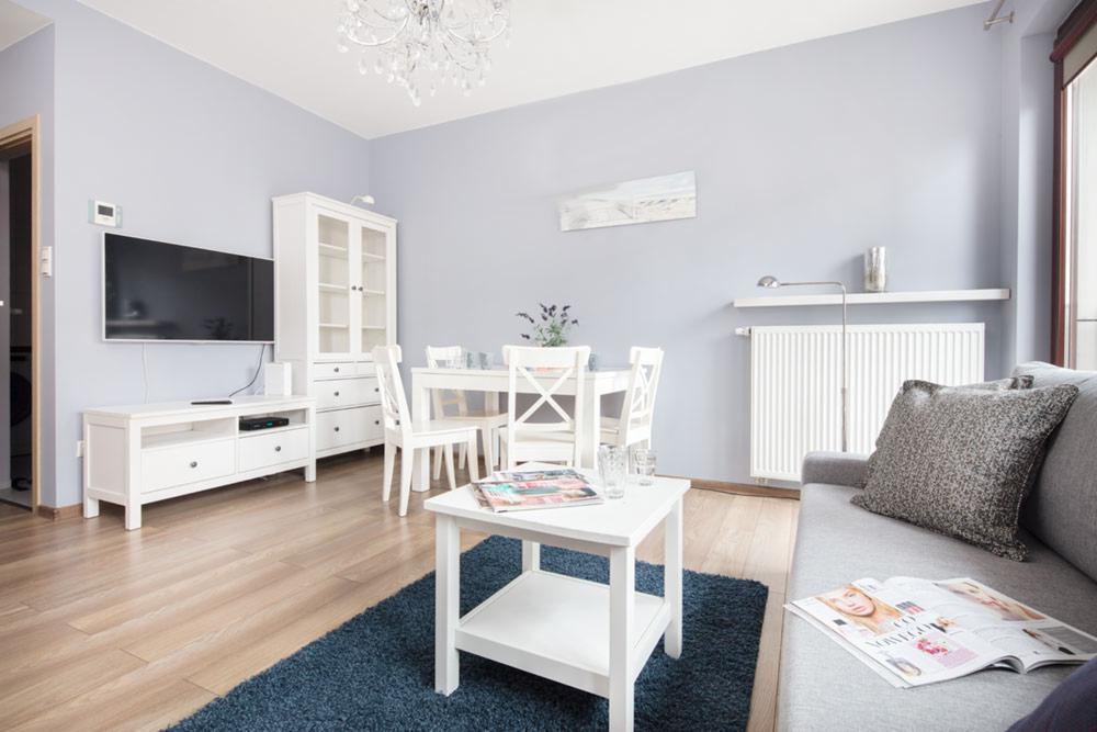 Jasne wnętrze salonu z białymi meblami, niebieskim dywanem i elementami dekoracyjnymi, podkreślone naturalnym światłem wpadającym przez okno.