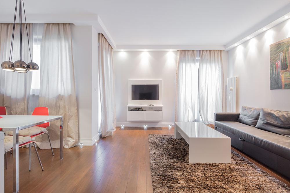 Jasne wnętrze salonu z drewnianą podłogą, białymi ścianami, szarą sofą, białym stolikiem kawowym na puszystym dywanie, telewizorem na ścianie, zasłonami i czerwonymi krzesłami przy białym stole.