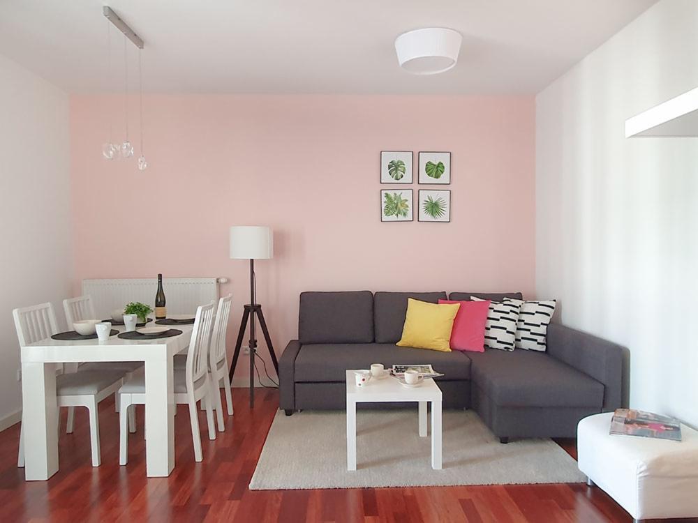 Jasne wnętrze mieszkania z różową ścianą, drewnianą podłogą, szarą sofą narożną z kolorowymi poduszkami, białym stołem z krzesłami i lampą na trójnogu.